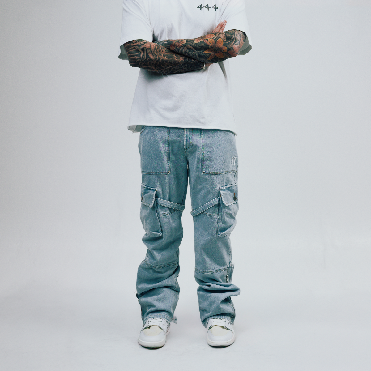 Person wearing light blue cargo pants and a white shirt with tattoos on arms against a plain background
